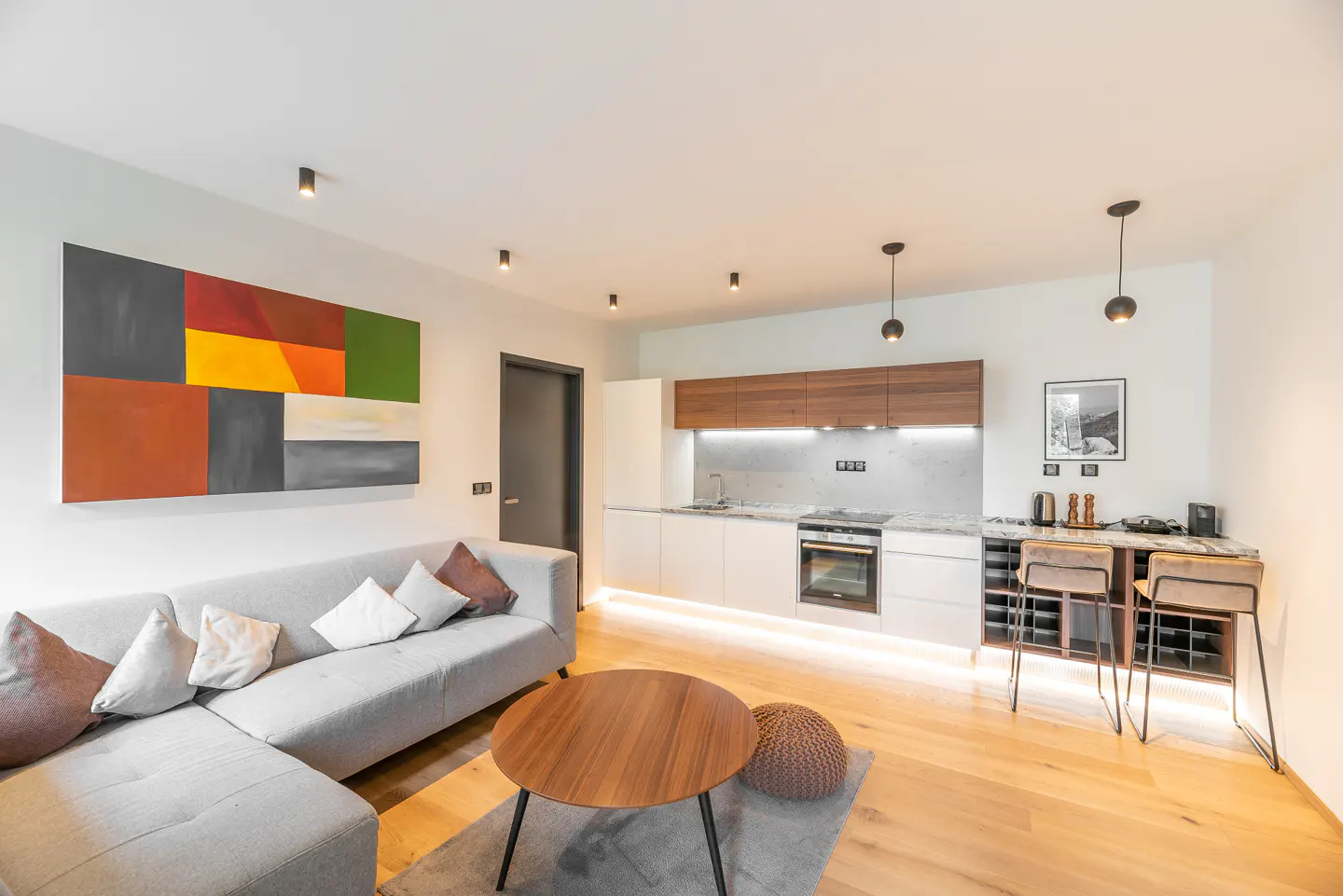 Open-concept living space with a gray sofa, colorful art, wood table, and a modern kitchen with bar seating.