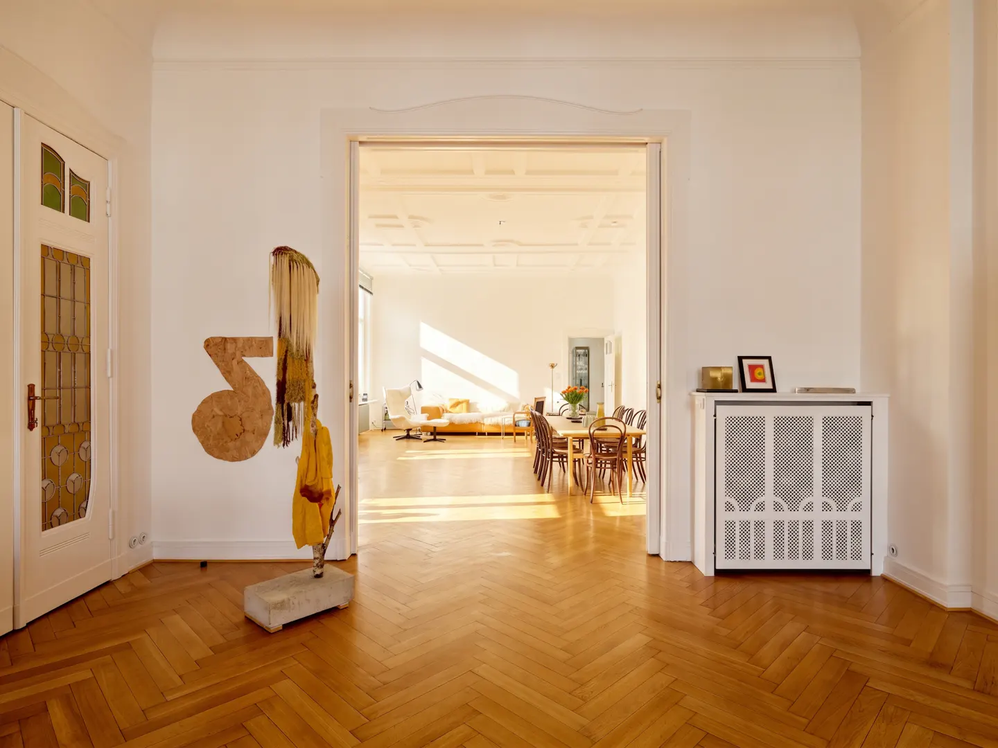 Bright, open-plan apartment with herringbone wood floors. A doorway leads to a dining area and living room with white walls and modern decor.