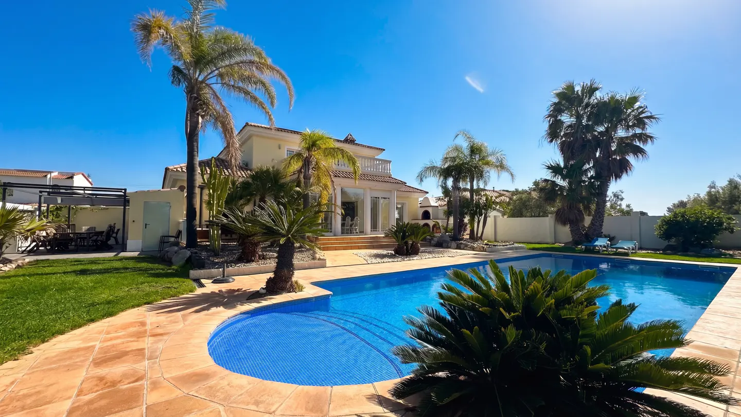 A sunny backyard with a blue tiled pool, palm trees, and a two-story yellow house with a balcony.