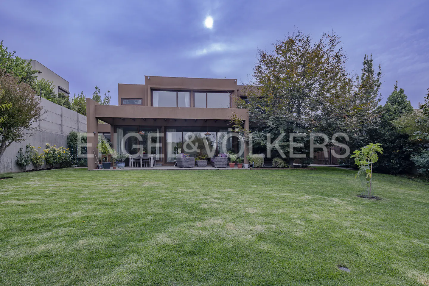 Two-story brown house with a large green lawn and trees under a cloudy sky. Patio furniture is visible under a covered area.