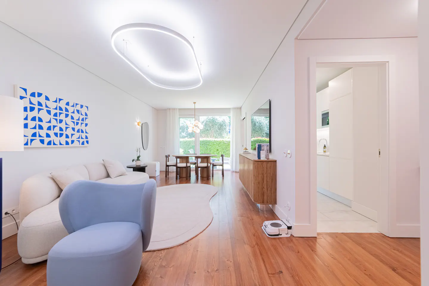 Bright, modern living room with white walls, wood floors, and a blue accent chair. A robot vacuum cleans near a wooden cabinet. Dining area visible through a window.