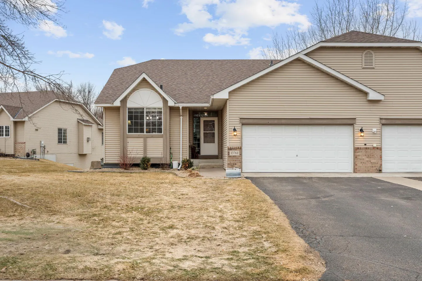 Beige two-story home with a brown roof, white garage door, and a lawn with dormant grass under a cloudy sky.