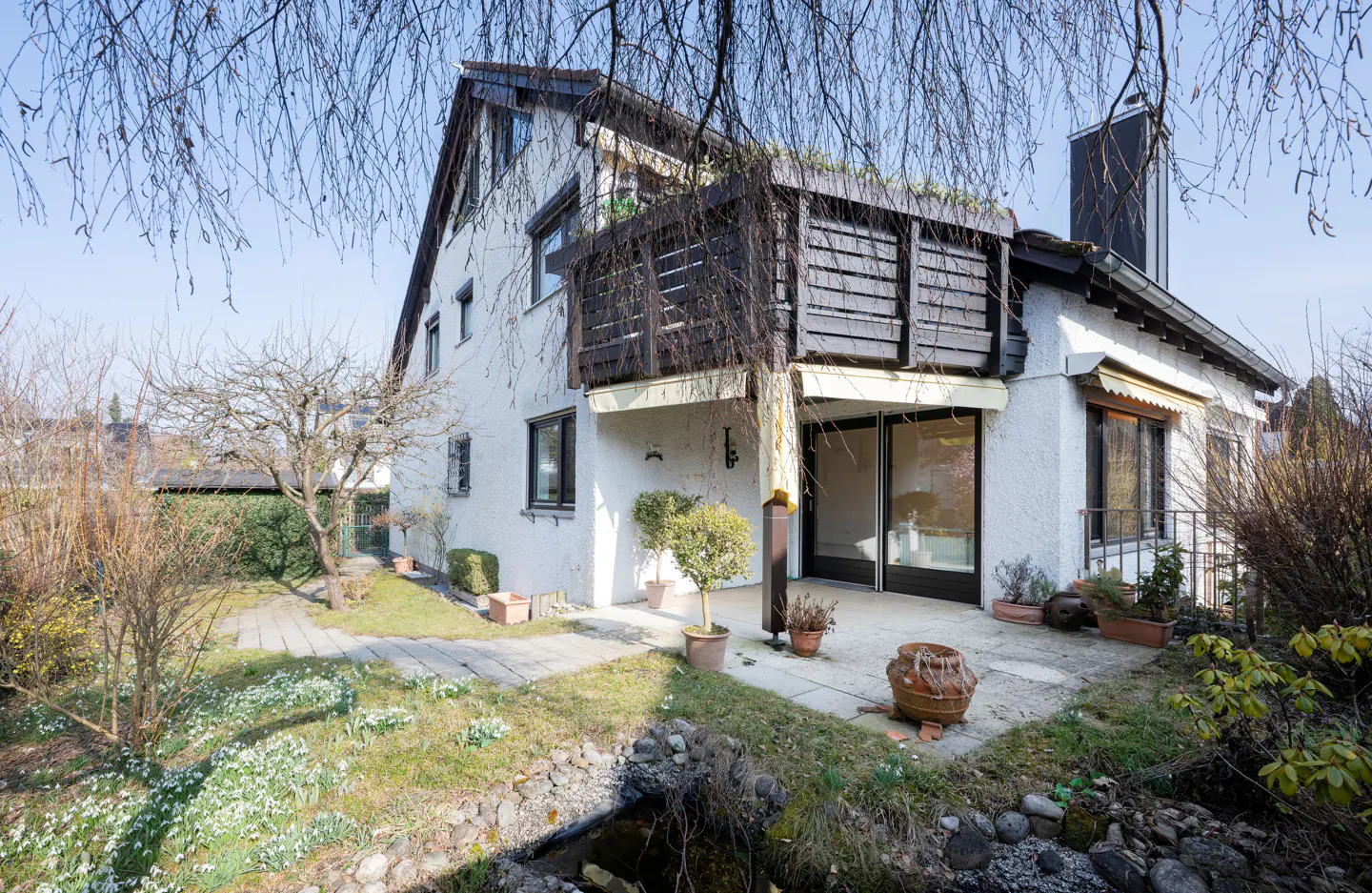 Exterior view of a two-story white house with a dark brown balcony and chimney, surrounded by a garden with a small stream.