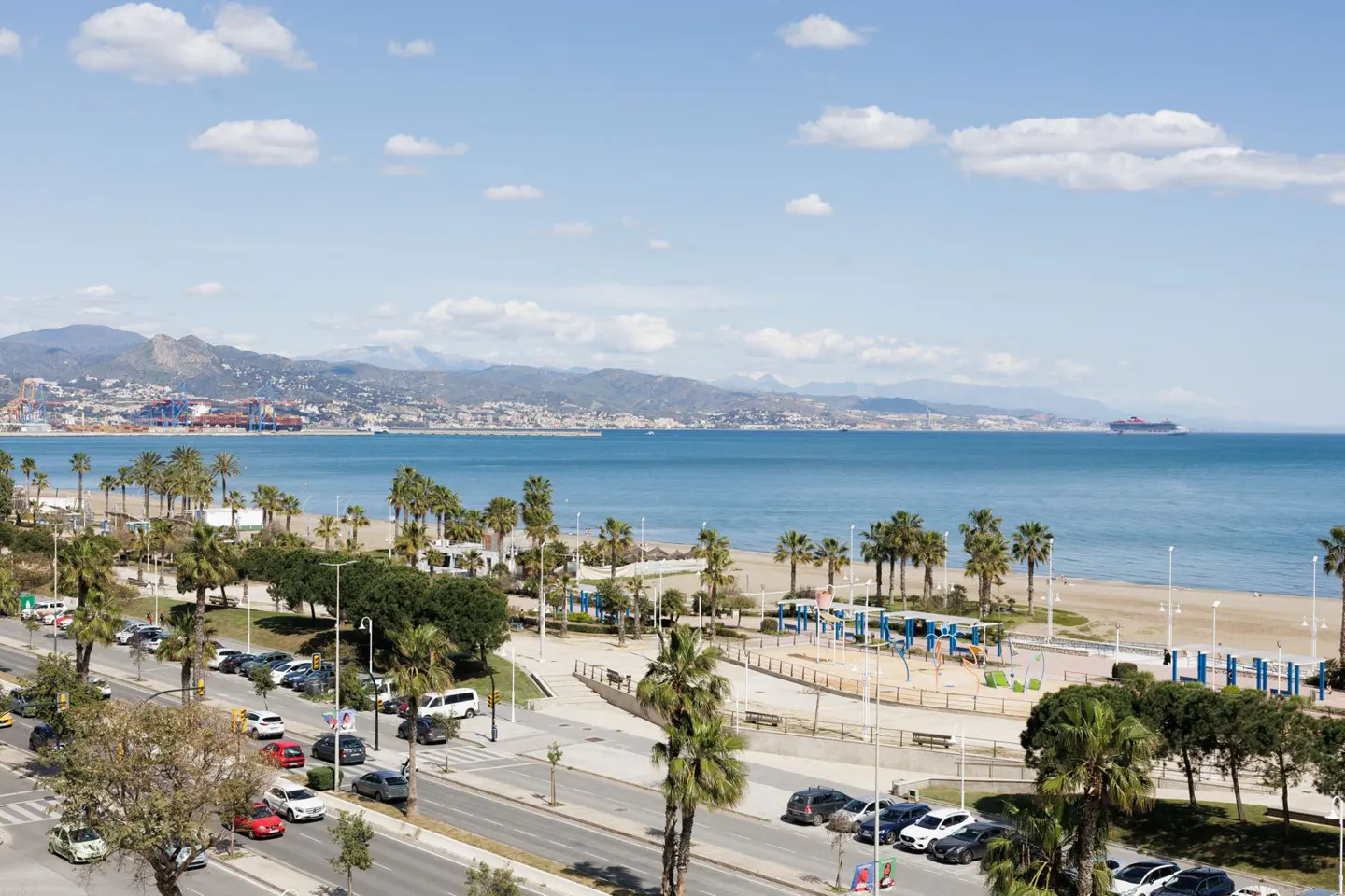 Scenic view of a beach with palm trees, a road with cars, and mountains in the background under a blue sky with clouds.