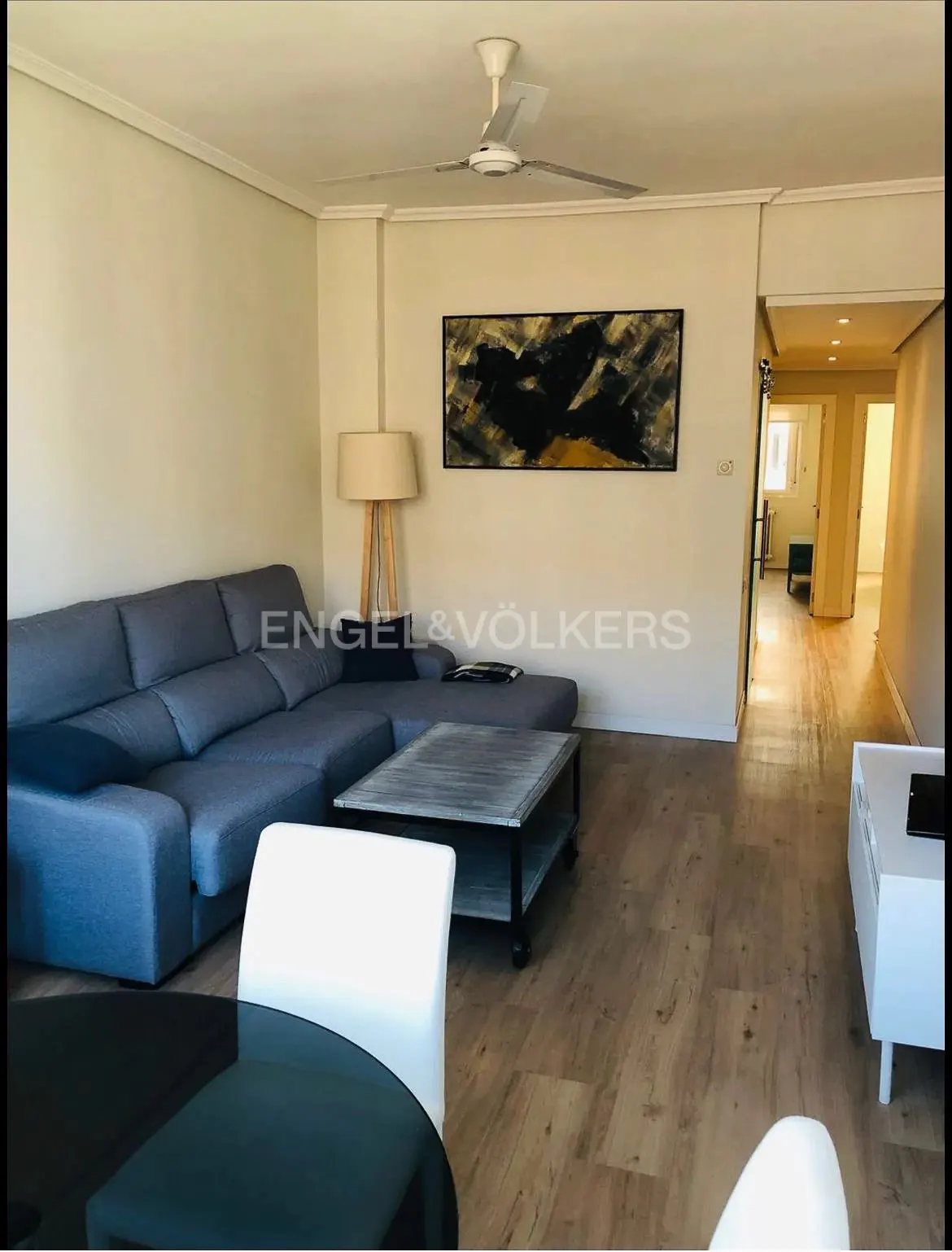 Living room with a gray sectional sofa, coffee table, and a black abstract painting on the wall. Hardwood floors and a ceiling fan are visible.