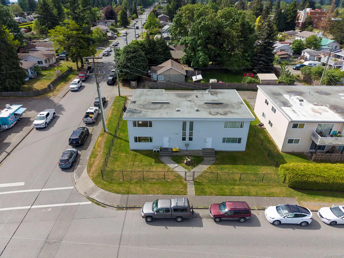 Aerial view of a light blue two-story duplex with a green lawn and black fence, cars parked on the street.