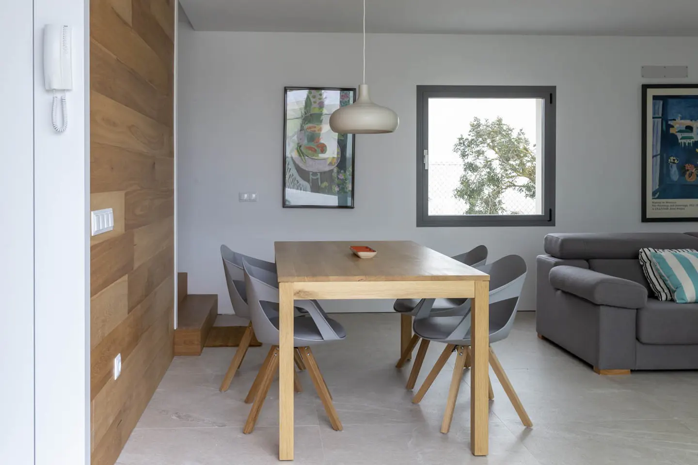 A modern dining area with a light wood table, gray chairs, and a gray sofa. A wood-paneled wall adds warmth to the white-walled space.