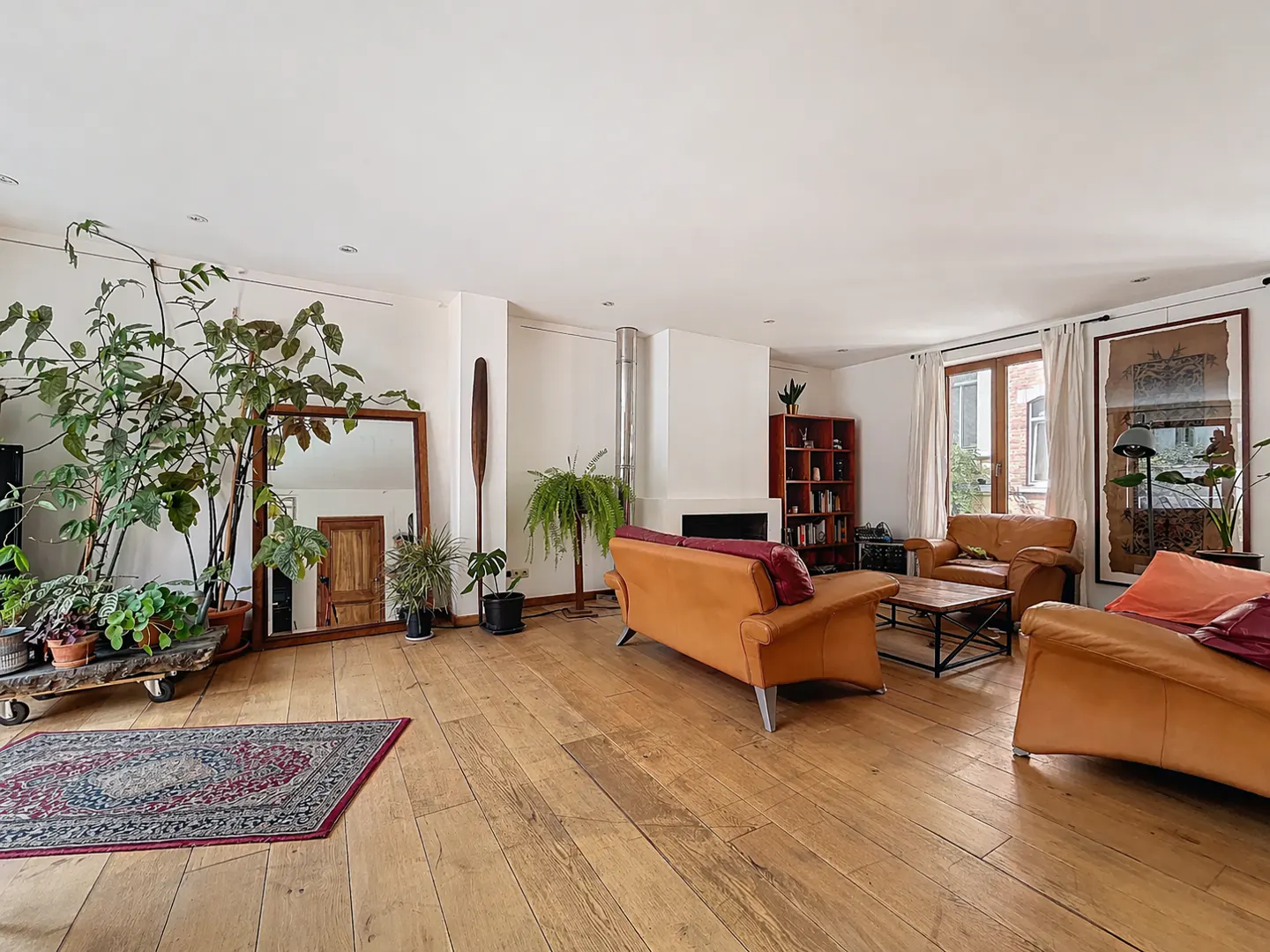 Bright living room with wood floors, white walls, and tan leather sofas. Plants, a mirror, and a fireplace add character.