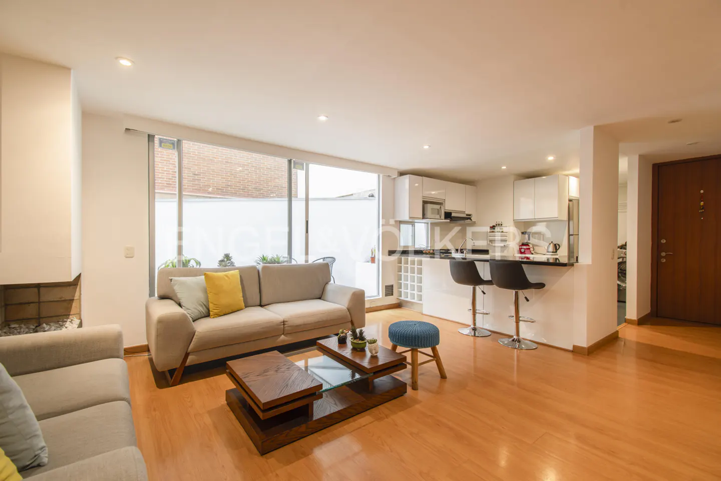 A bright, open-concept living space with a beige sofa, wood floors, and a modern white kitchen. A large window provides natural light.