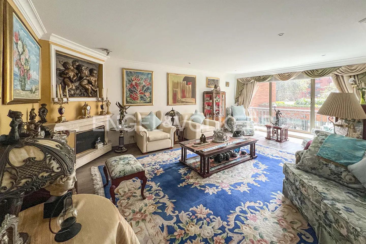 A living room with beige chairs, a blue floral rug, and art on the walls. A fireplace is visible.
