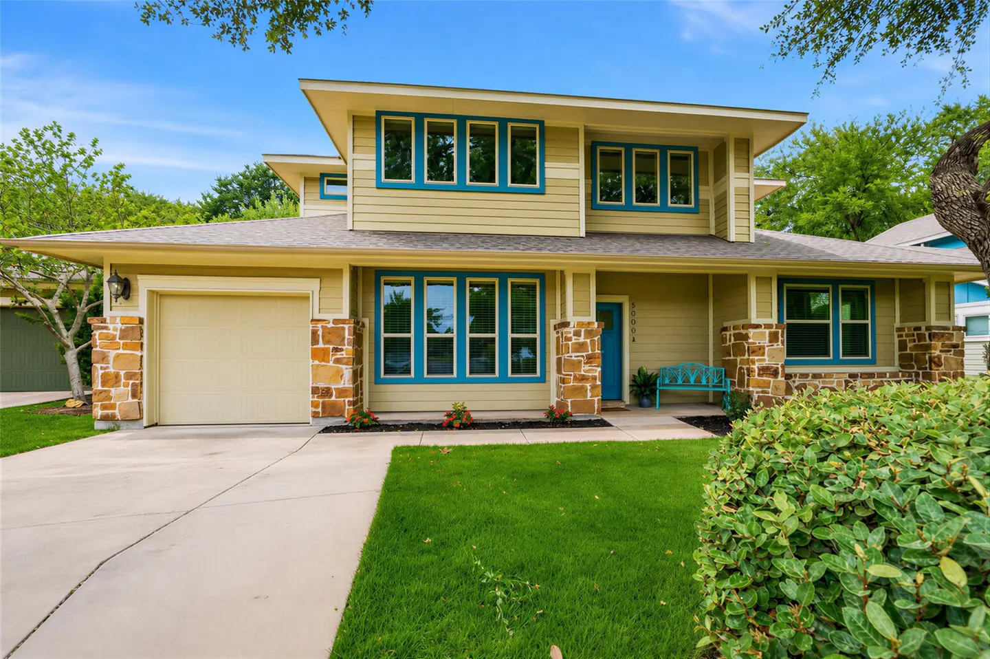 Two-story tan house with stone accents, blue window trim, a blue front door, a garage, and a green front lawn.