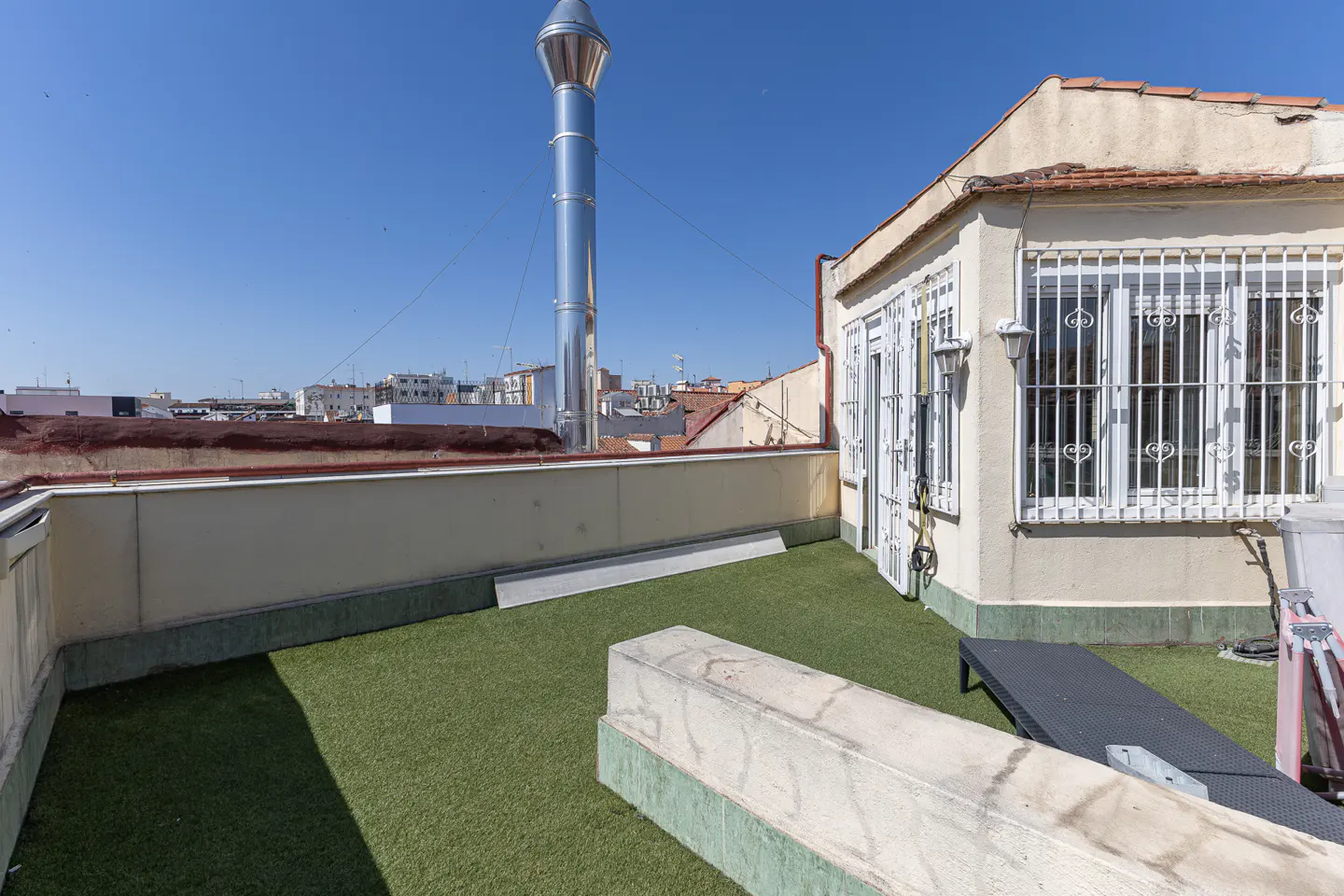 Rooftop terrace with artificial grass, a low stone wall, a metal chimney, and a building exterior with barred windows.