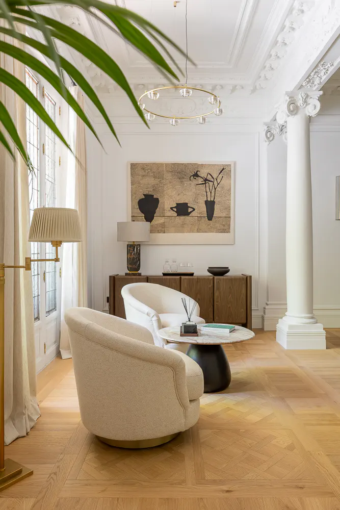Bright living room with parquet floors, white walls, and ornate ceiling. Two cream-colored chairs face a round marble table. A large column stands near a wooden cabinet with art above.