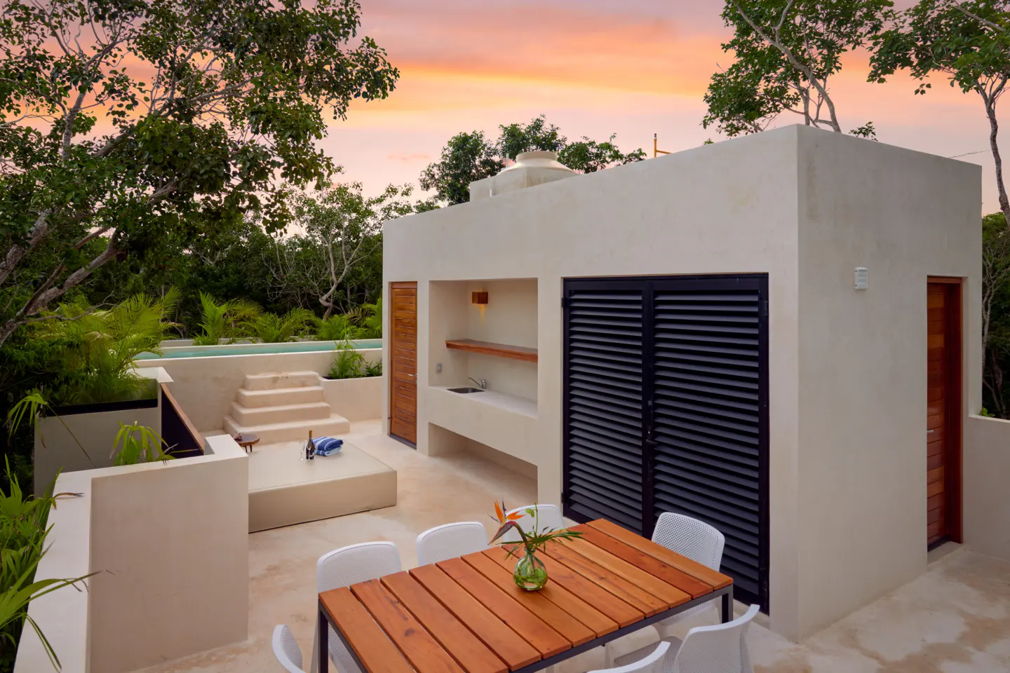 Modern outdoor patio with a wooden dining table, kitchenette, and small pool at sunset surrounded by lush greenery.