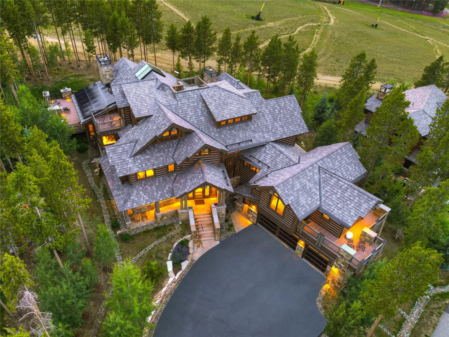 Aerial view of a large, luxurious log cabin home with a gray shingle roof, surrounded by green trees and a grassy hill.