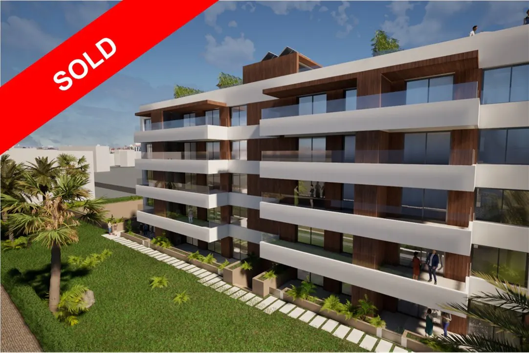 A modern, multi-story apartment building with white balconies and wood accents, marked "SOLD" in red. Lush green lawn and palm trees surround the building.