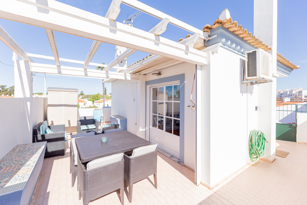 Outdoor patio with a white pergola, table, chairs, and a bench with colorful tiles. An air conditioner and a green garden hose are on the white wall.