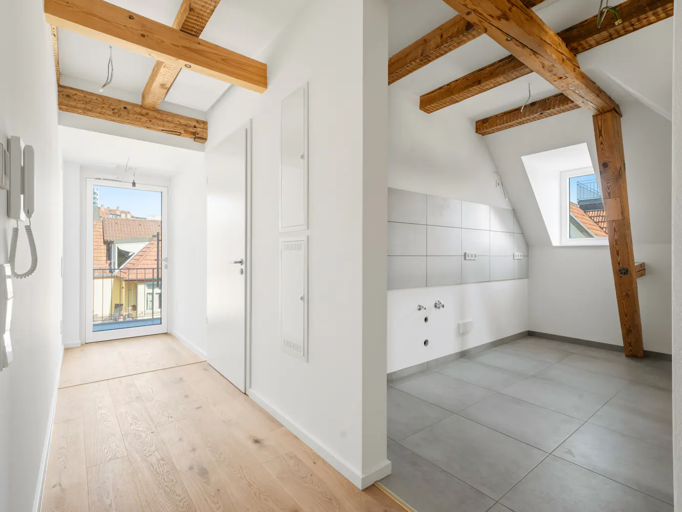 Bright, modern apartment interior with exposed wooden beams, white walls, and light wood floors leading to a balcony. A kitchen area features gray tile.