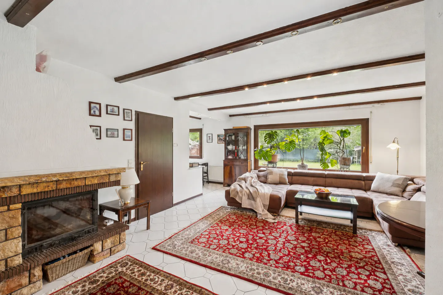 Living room with a brick fireplace, brown leather sectional sofa, and a red patterned rug. A large window overlooks a green garden.