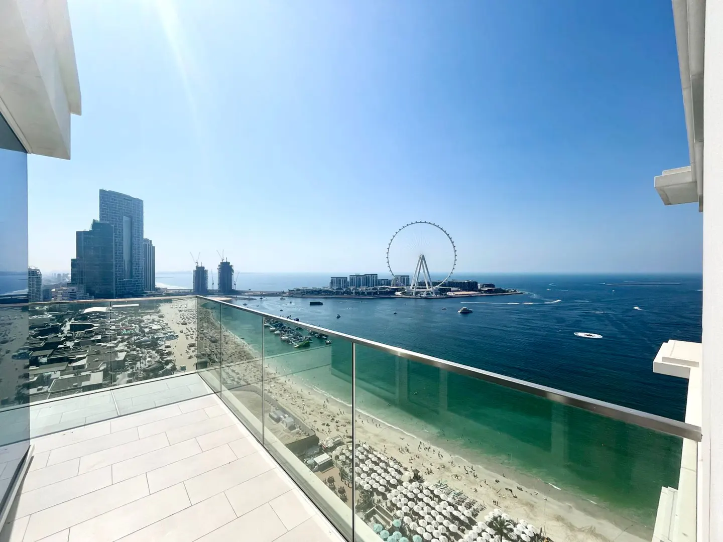Balcony view of Dubai Eye, beach, and ocean. Glass railing, white tile floor, blue sky.