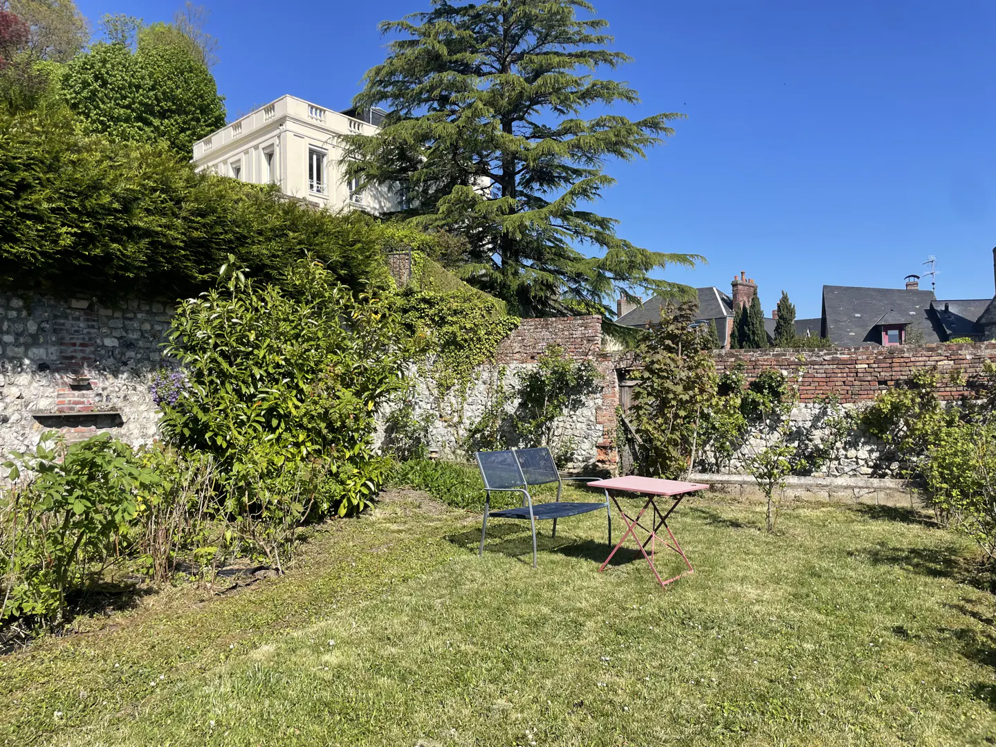 A garden with a metal chair and table on the grass, a brick wall, and a house in the background.