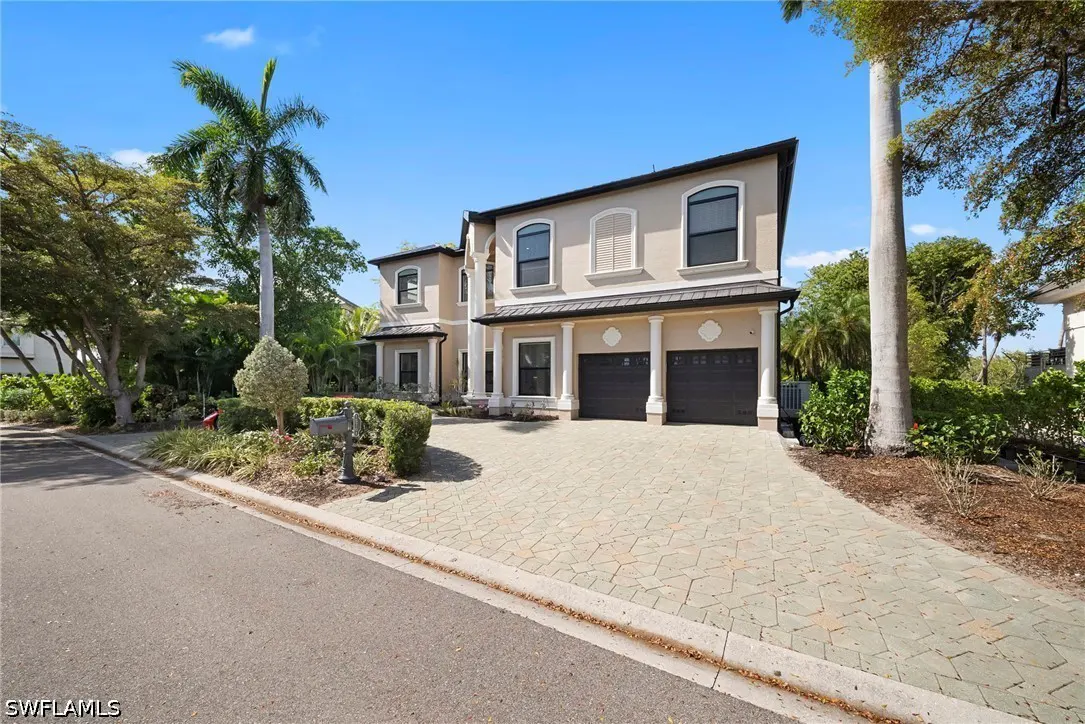 Two-story beige house with black trim, two-car garage, and a patterned driveway under a blue sky. Palm trees and greenery surround the property.