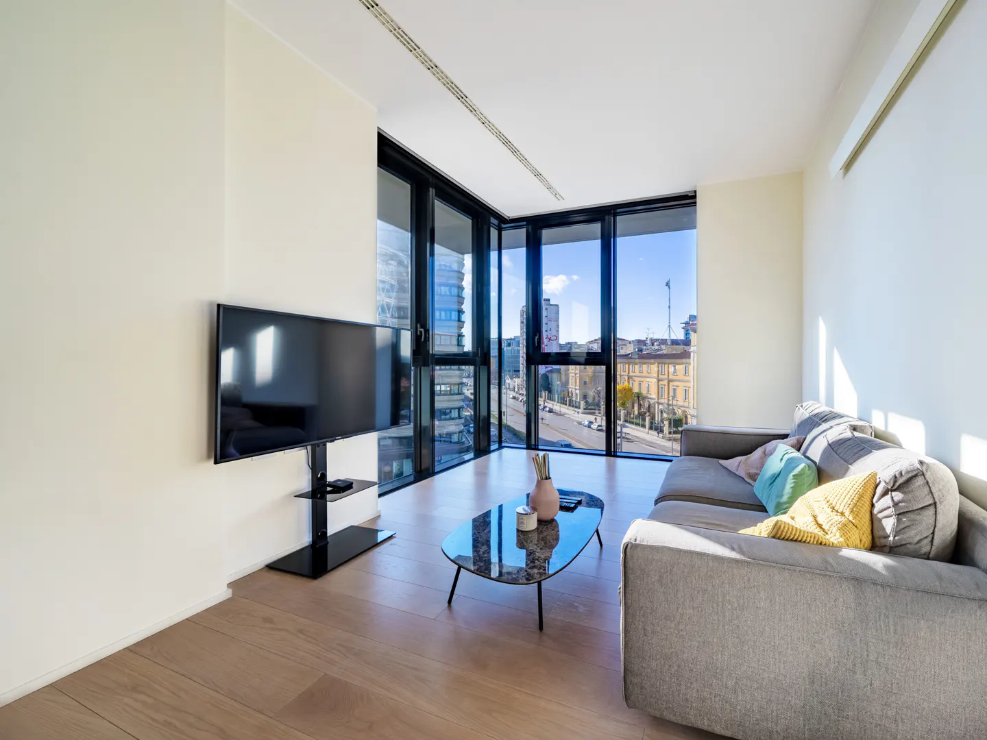 Bright living room with wood floors, gray sofa with colorful pillows, and a black TV against a large window with a city view.