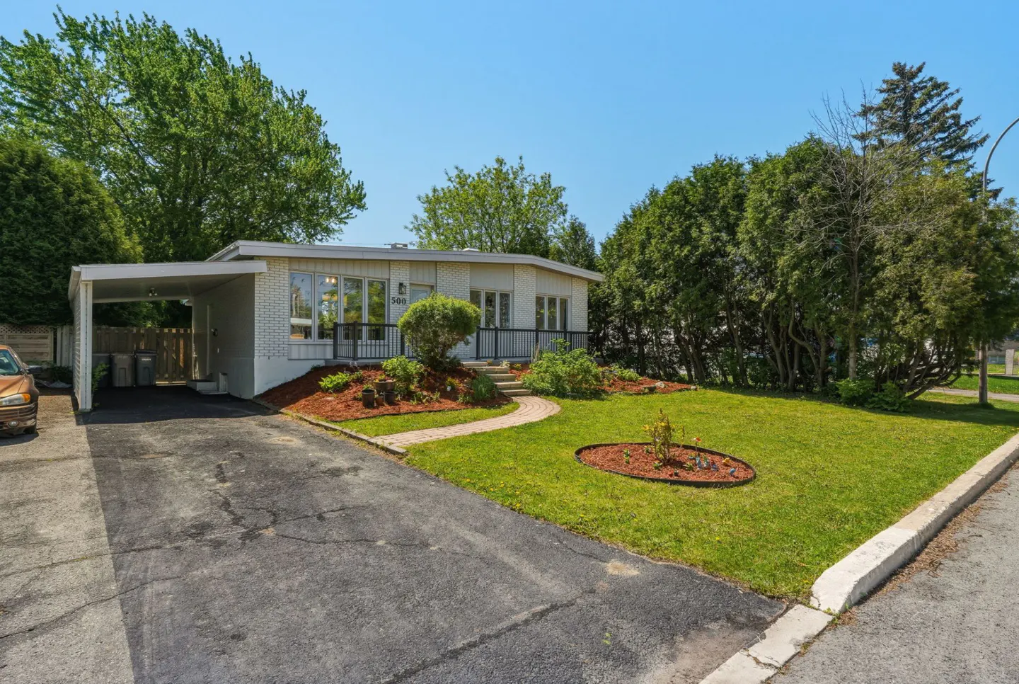 Single-story white brick house with a carport, paved driveway, and a landscaped front yard under a clear blue sky.