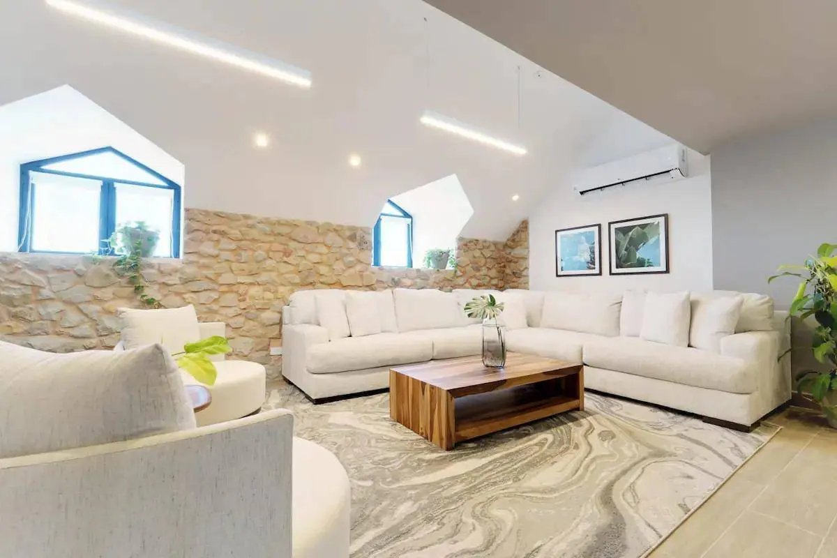Bright living room with stone wall, white sofa, and wood coffee table. Windows with blue frames and plants add natural light.