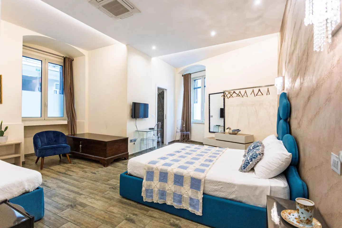 Bright hotel room with a blue bed frame, white bedding, and a blue and white quilt. There is a blue chair and a wooden chest in the room.