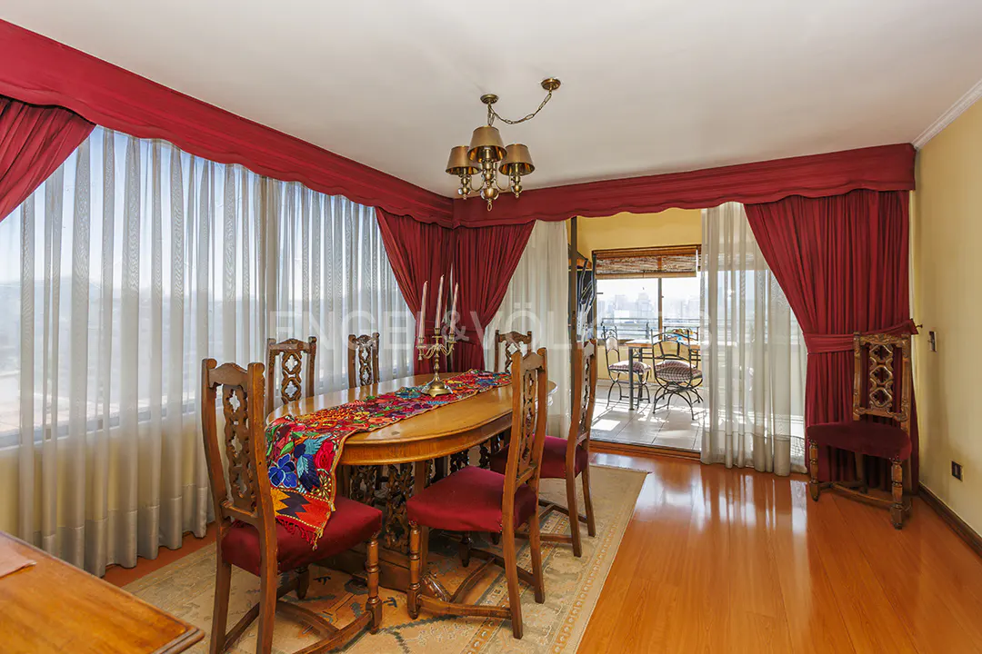 Dining room with wood table, red chairs, and a colorful runner. Red curtains frame sheer windows and a balcony with outdoor seating.