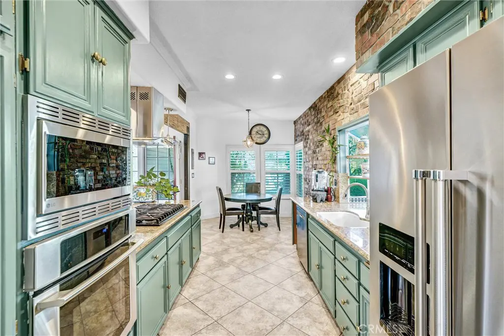 A kitchen with light green cabinets, stainless steel appliances, and a brick accent wall. A dining table is visible in the background.