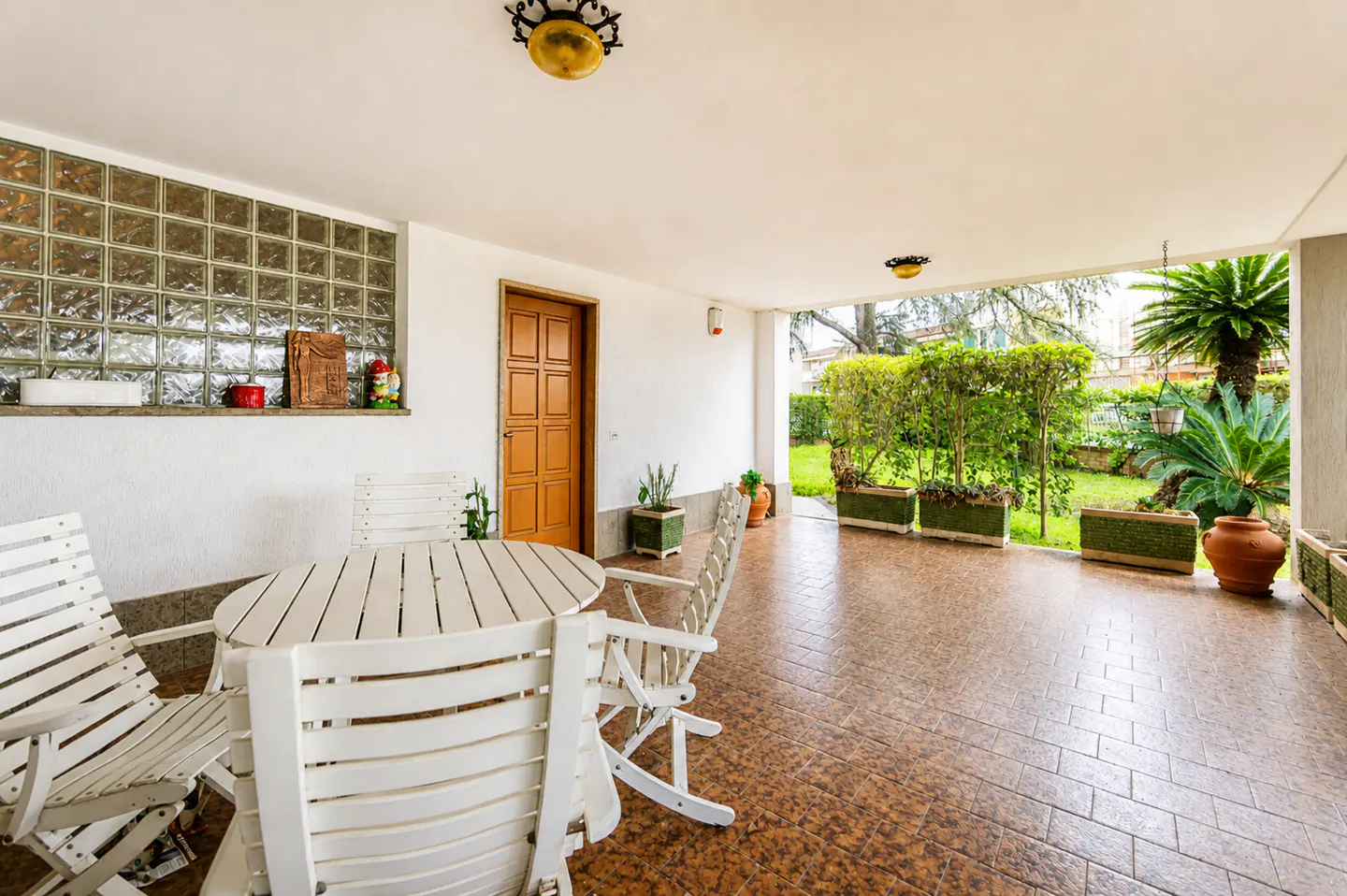 Covered patio with white table and chairs, brown tile floor, and a view of a green garden.