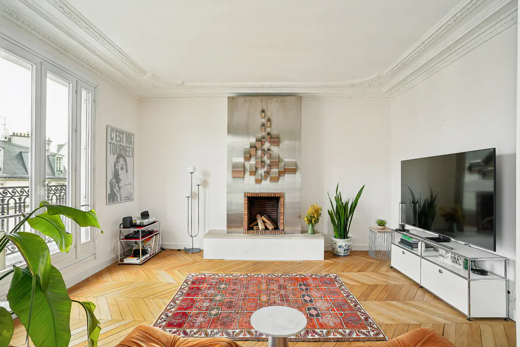 Bright living room with herringbone floors, a red patterned rug, and a modern fireplace with a silver accent wall. A large window lets in natural light.