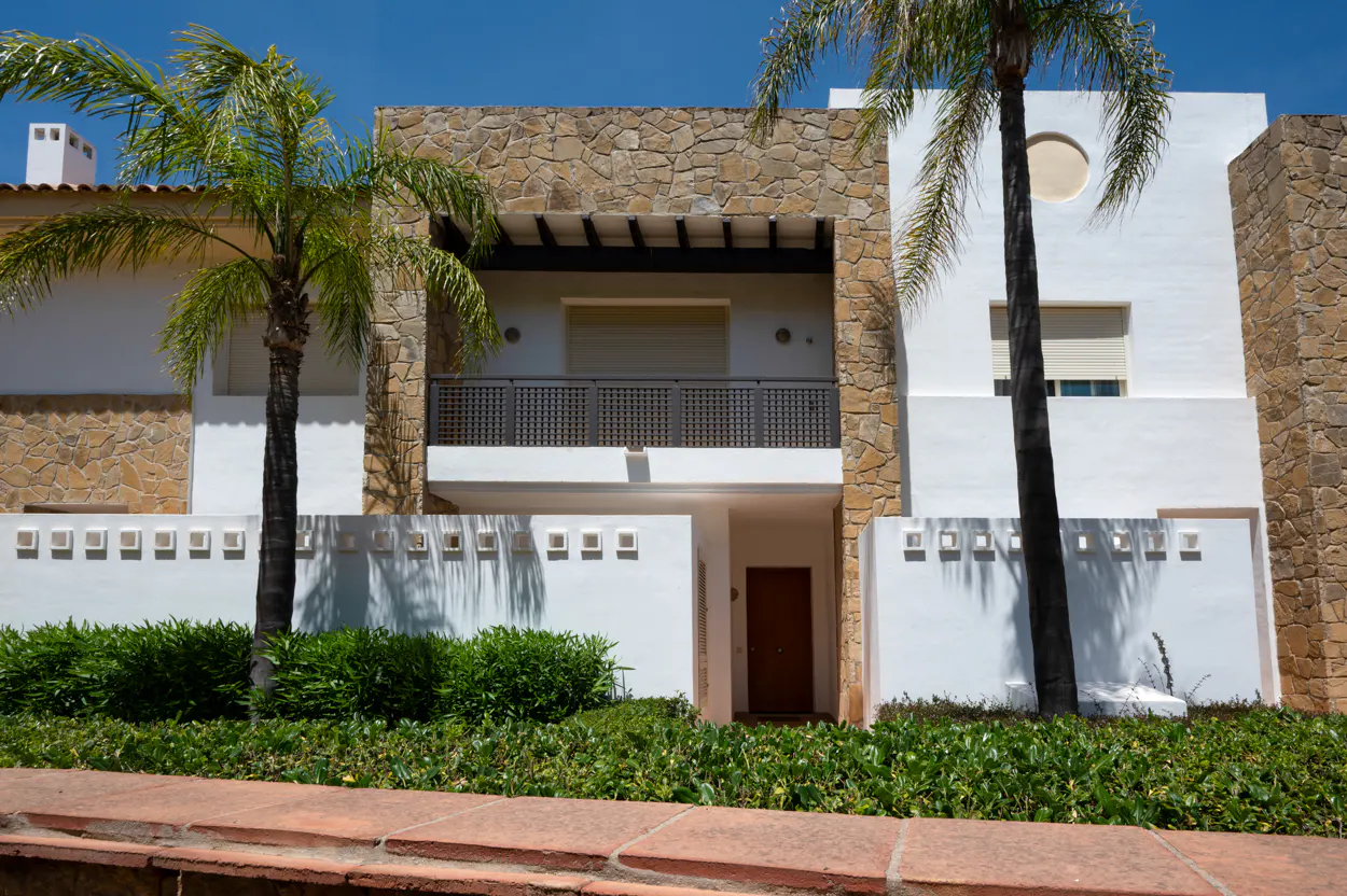 Modern two-story house with white walls, stone accents, balcony, and palm trees under a blue sky.