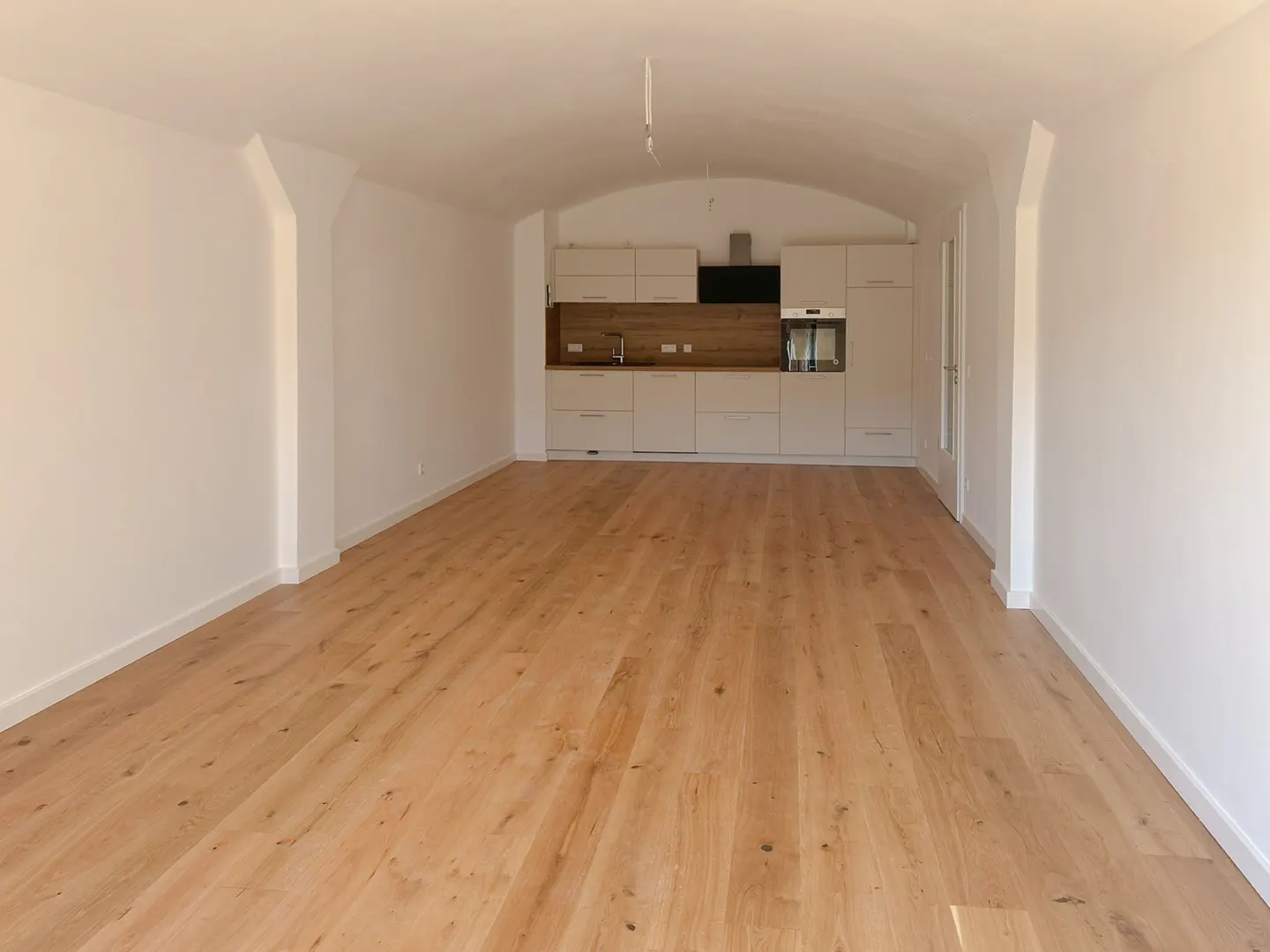Bright, empty room with light wood floors and white walls. A modern kitchen is visible in the background.