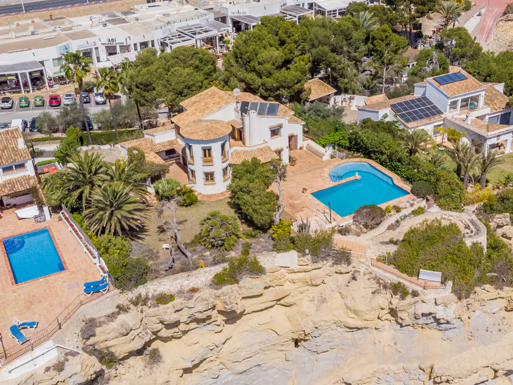 Aerial view of a white house with a pool, terracotta roof, and a cliffside location. Lush greenery surrounds the property.