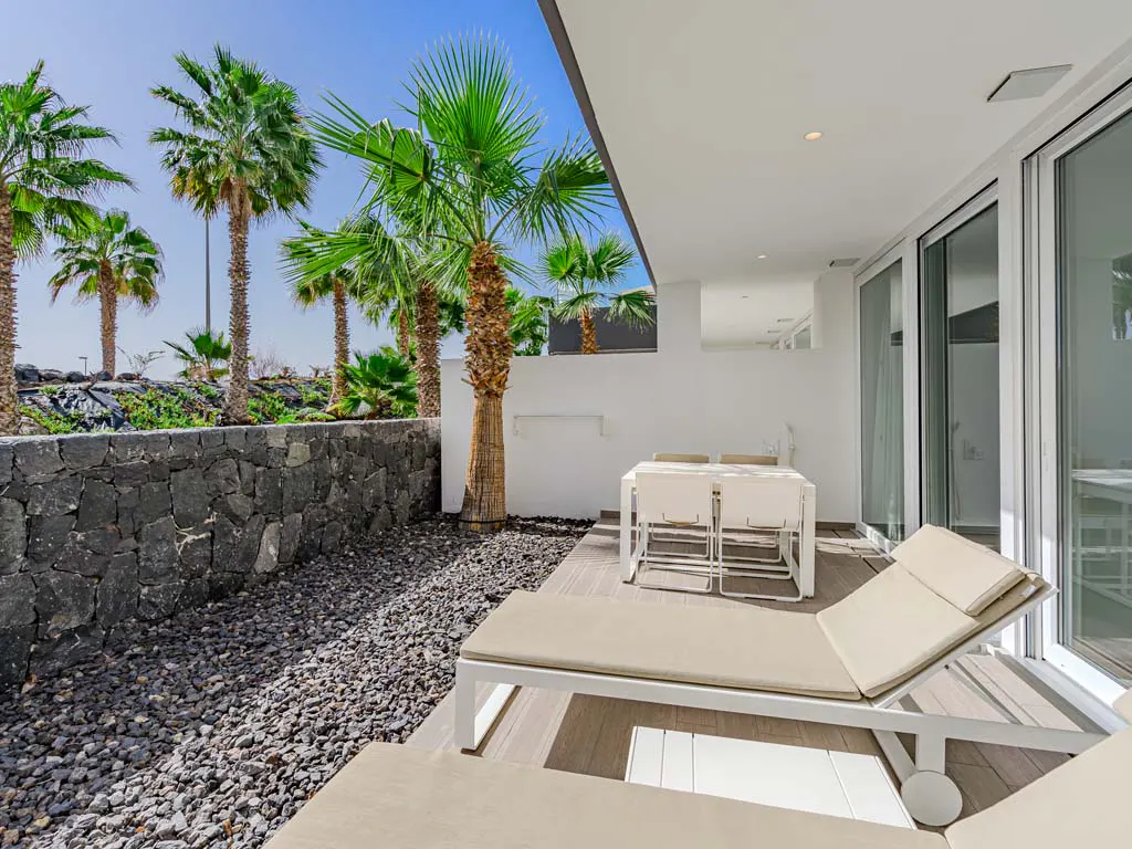 Outdoor patio with white furniture, including a table, chairs, and lounge chairs. Palm trees and a stone wall are visible in the background.