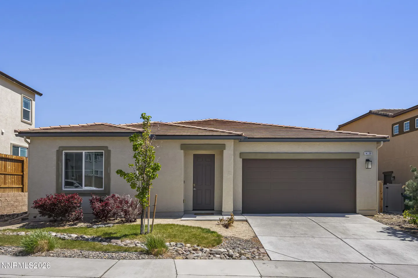 Beige single-story home with a brown roof and garage door under a clear blue sky. Landscaped front yard with small tree and bushes.