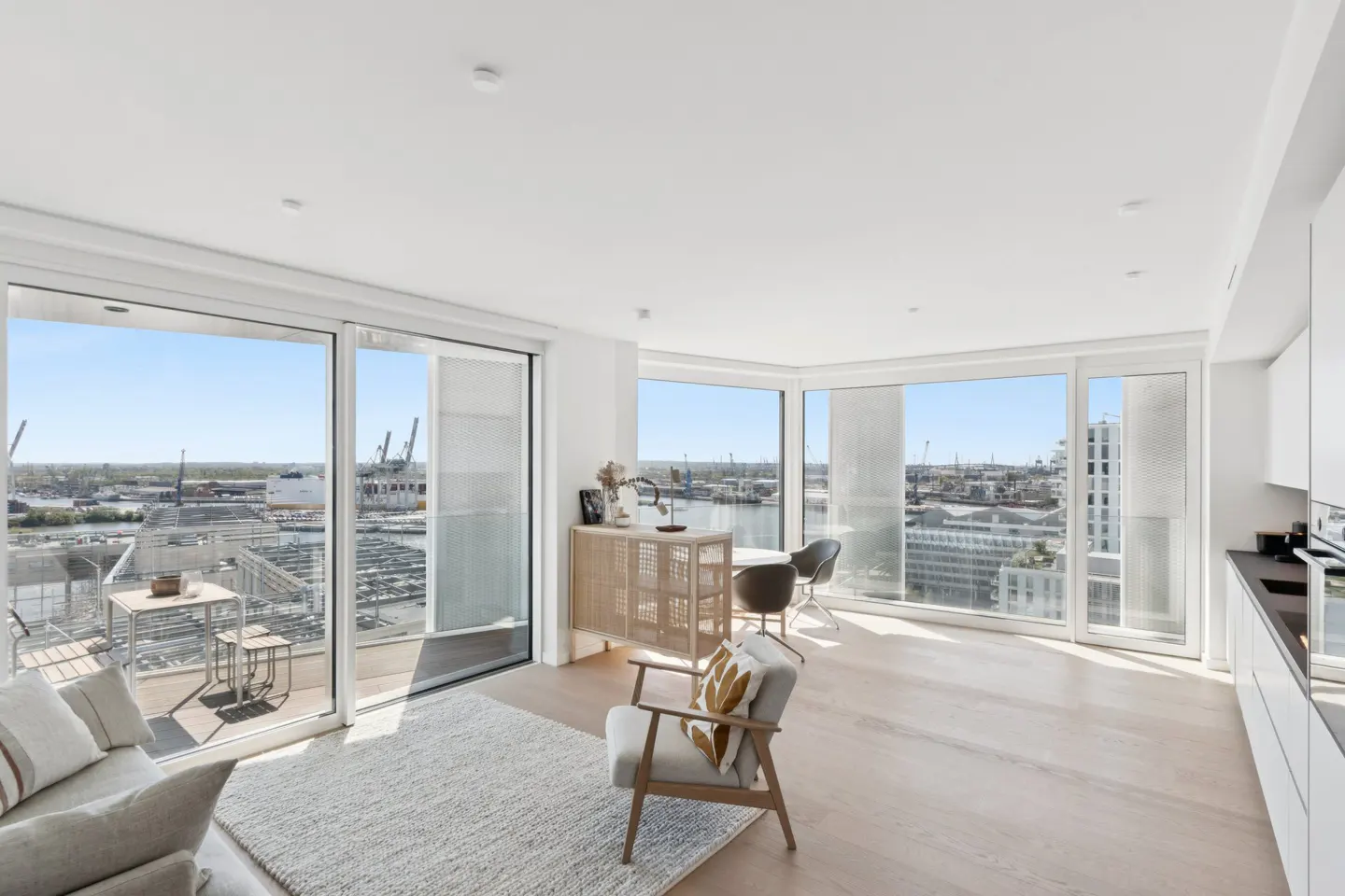 Bright, modern apartment interior with large windows overlooking a harbor. A gray sofa, chair, and dining set furnish the space.