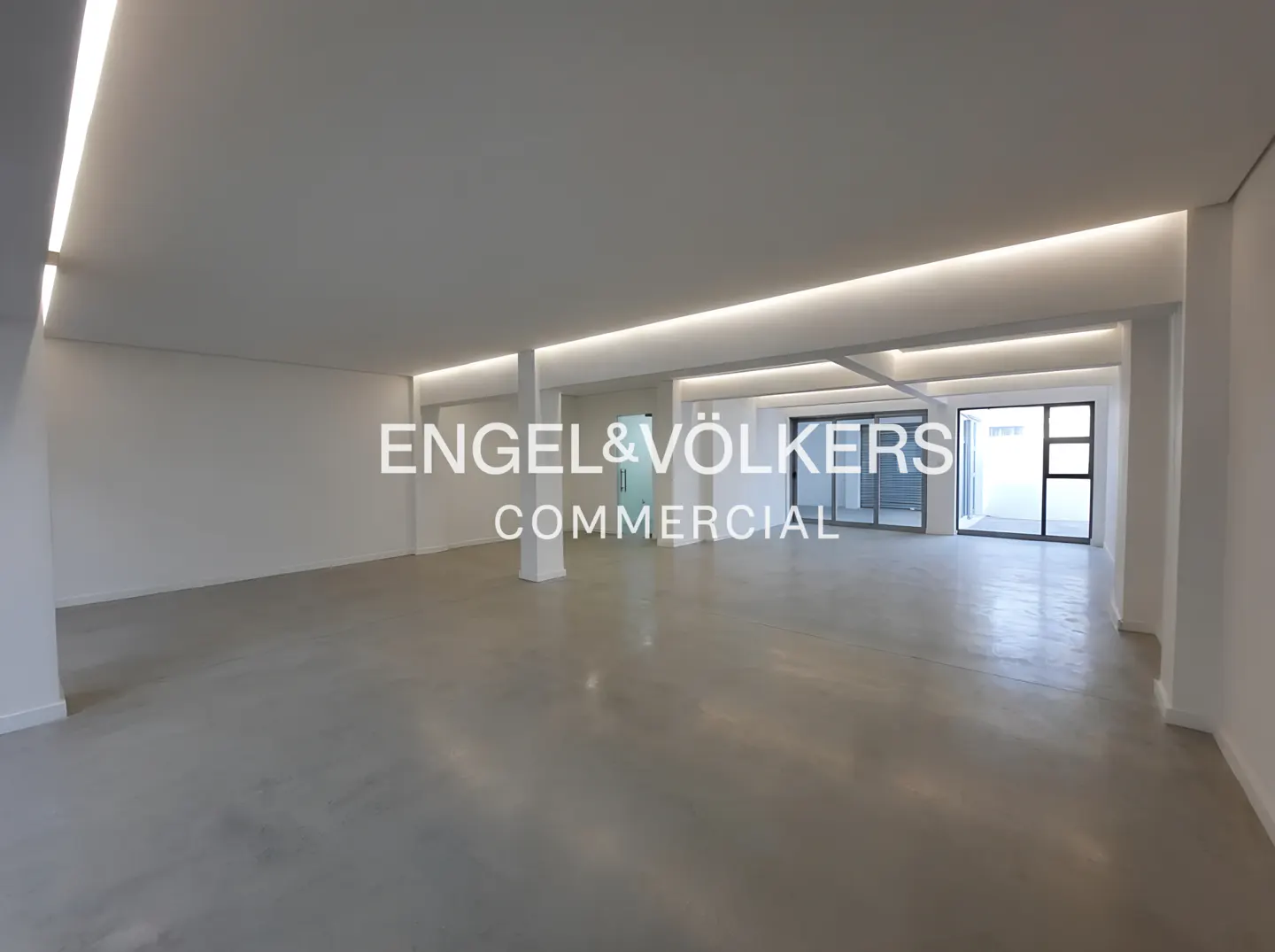 Empty commercial space with white walls, gray floor, and recessed lighting. Glass doors lead to an outdoor area.