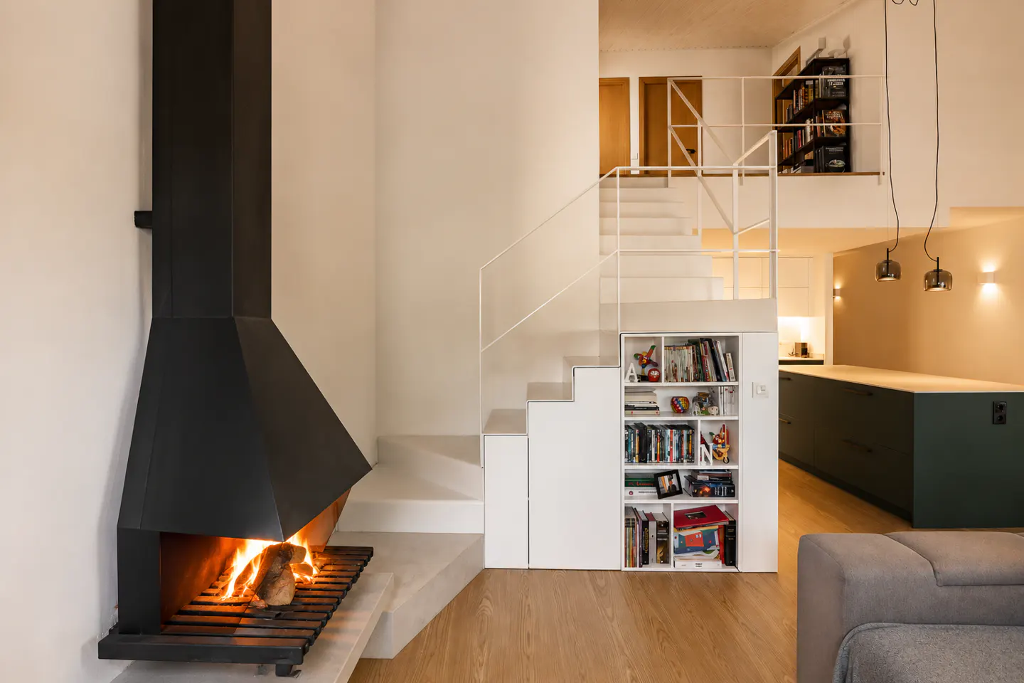 Modern living room with a black fireplace, white staircase featuring built-in shelving, and a view of a kitchen area.