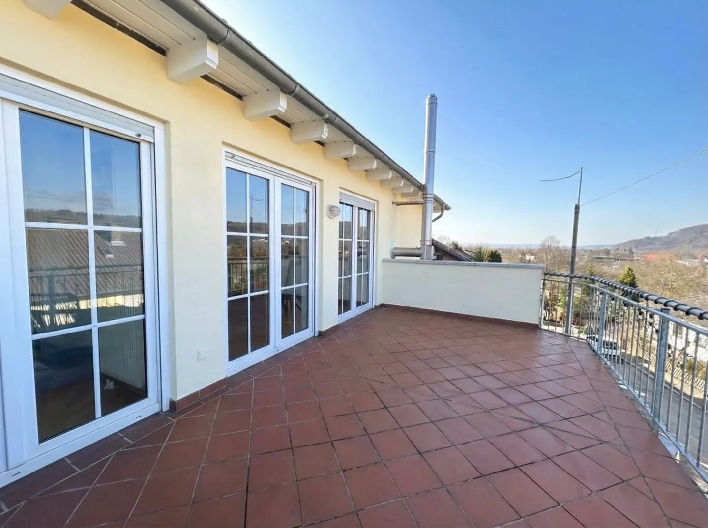 A sunny balcony with red tile flooring, white framed doors, and a metal railing overlooking a distant landscape.