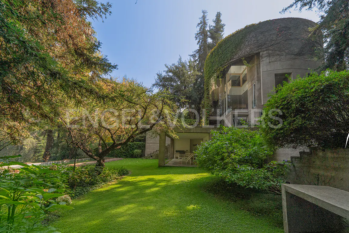 Exterior view of a modern, cylindrical home covered in ivy, surrounded by lush green trees and lawn.