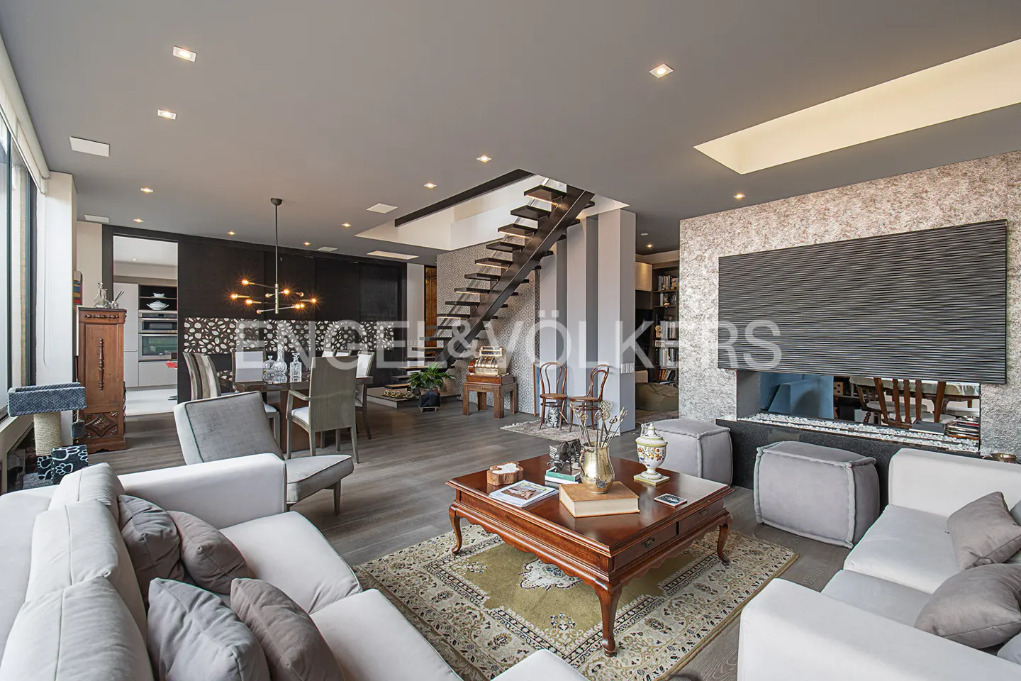 Open-concept living room with white sofas, wood coffee table on a patterned rug, and a modern fireplace with a large, dark textured panel.