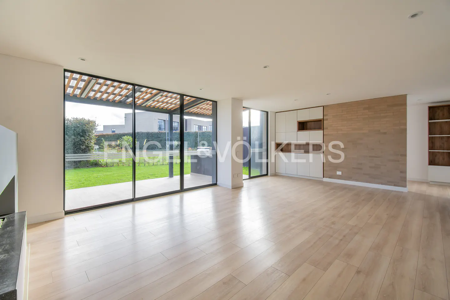Bright, empty living room with light wood floors, white cabinets, and large windows overlooking a green lawn.