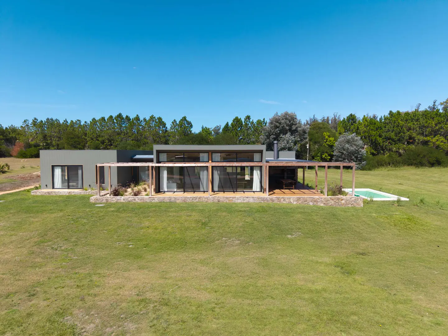Modern gray house with large windows and wood pergola on a green lawn, with trees in the background and a small pool.