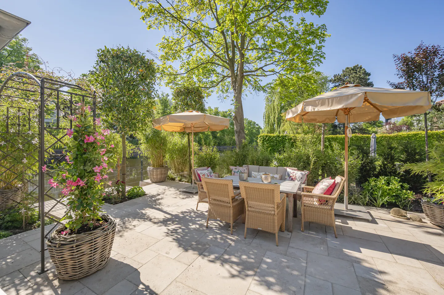 A sunny stone patio features a wicker dining set under beige umbrellas, surrounded by lush greenery and a metal trellis.