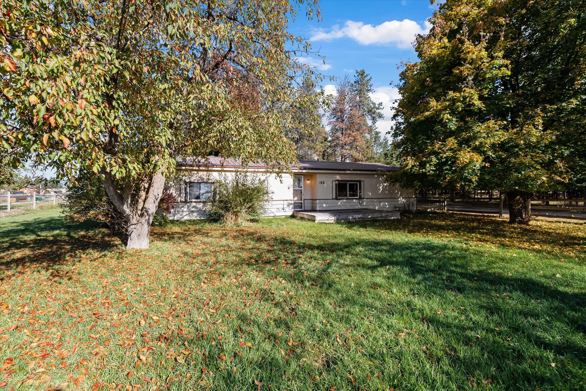 Single-story beige home with a front porch, surrounded by large trees and a lawn covered in autumn leaves under a blue sky. [AI-generated text]