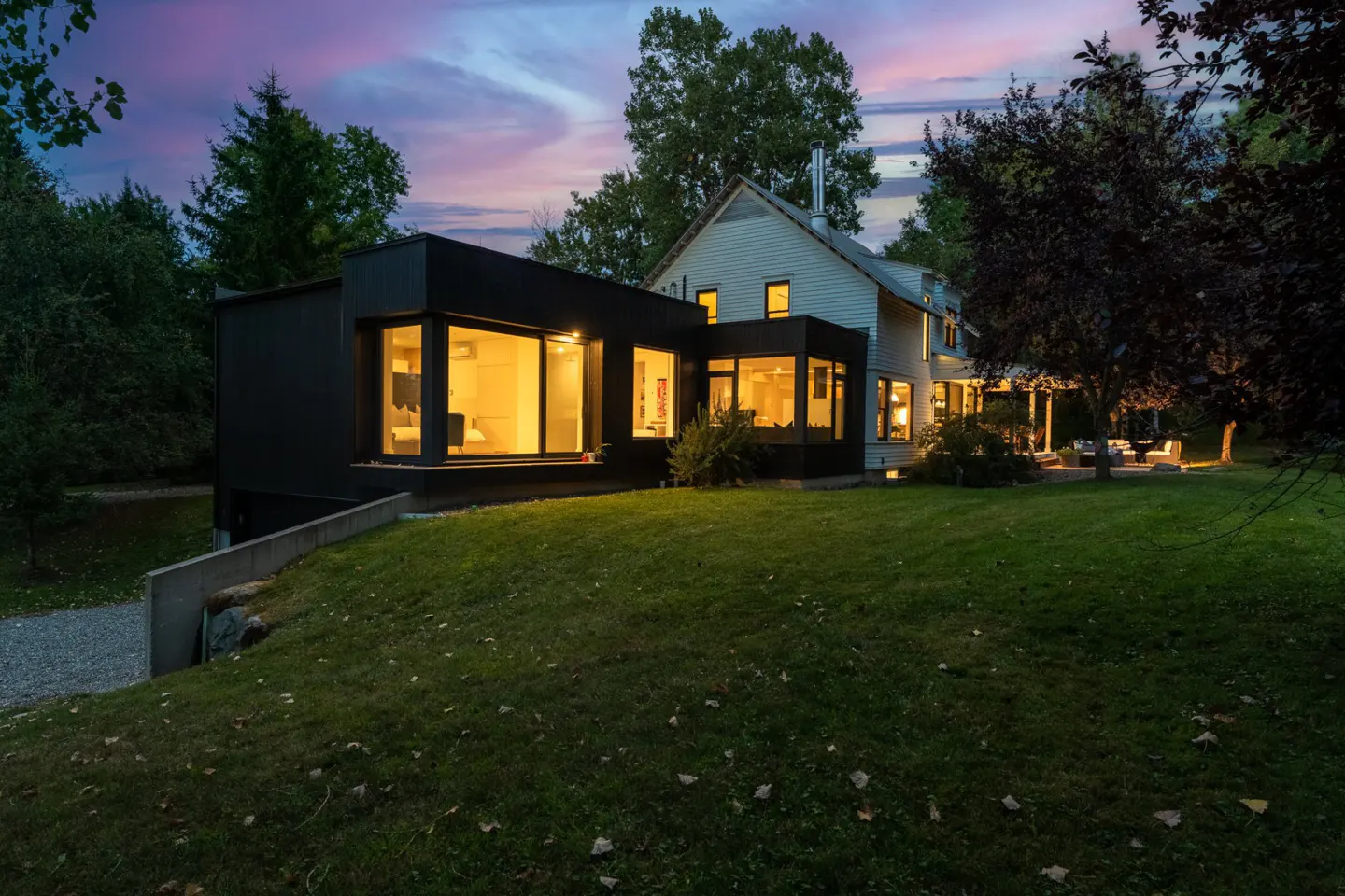 Twilight view of a modern farmhouse with a black addition, set on a green lawn with trees. Interior lights glow warmly.